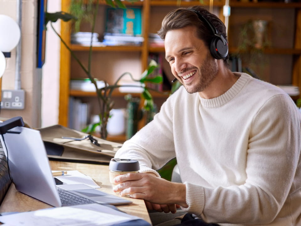 Evolve2 65 Flex wireless over-ear headset worn by a person working at a laptop and holding a coffee — home office setup.