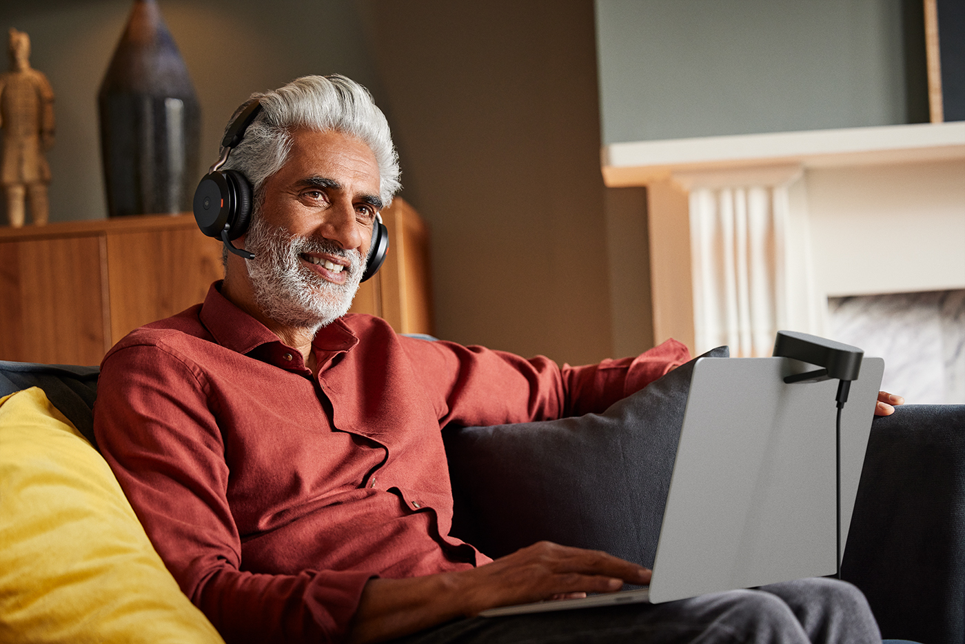 Man sitting comfortably on a couch at home, wearing a Jabra Evolve2 75 headset and using a laptop equipped with a Jabra PanaCast 20 camera, smiling during a video call.